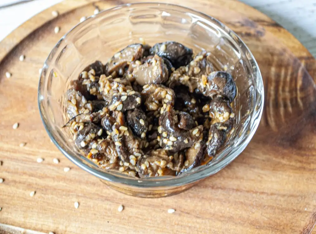 A glass bowl filled with marinated shiitake mushrooms. The bowl sits on a round wooden platter.