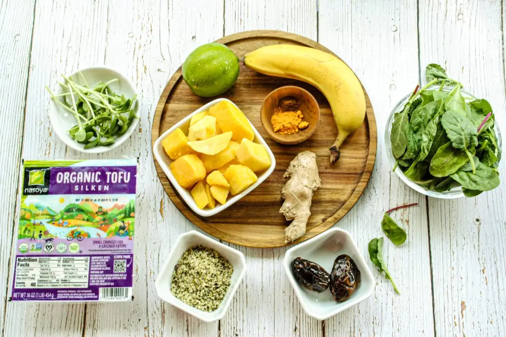 Smoothie ingredients on a table. Includes spinach, banana, a lime, mango, ginger, spices, silken tofu, dates and microgreens.