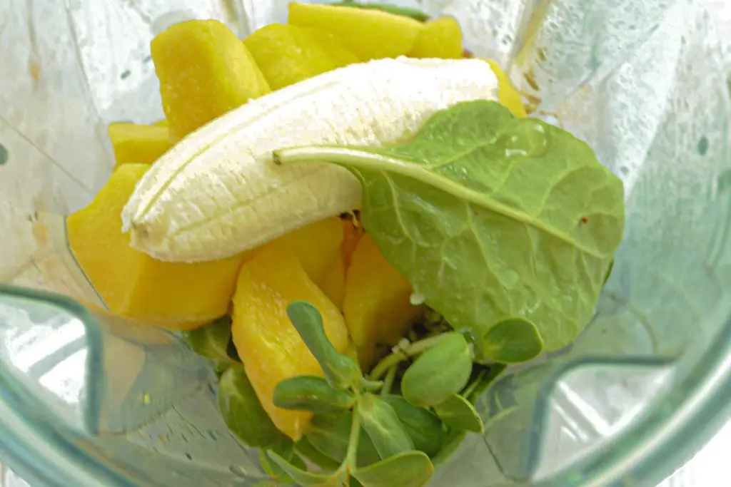 A blender carafe filled with mango chunks, a peeled banana and spinach.