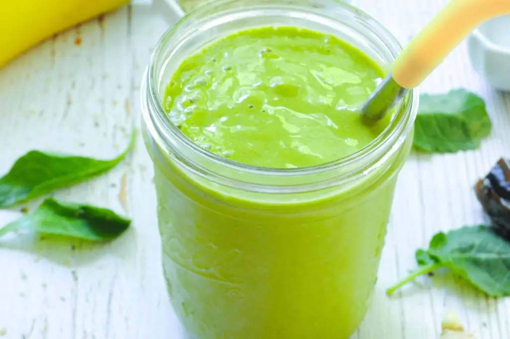 A green smoothie in a glass with a metal straw. It sits on a white wooden table.