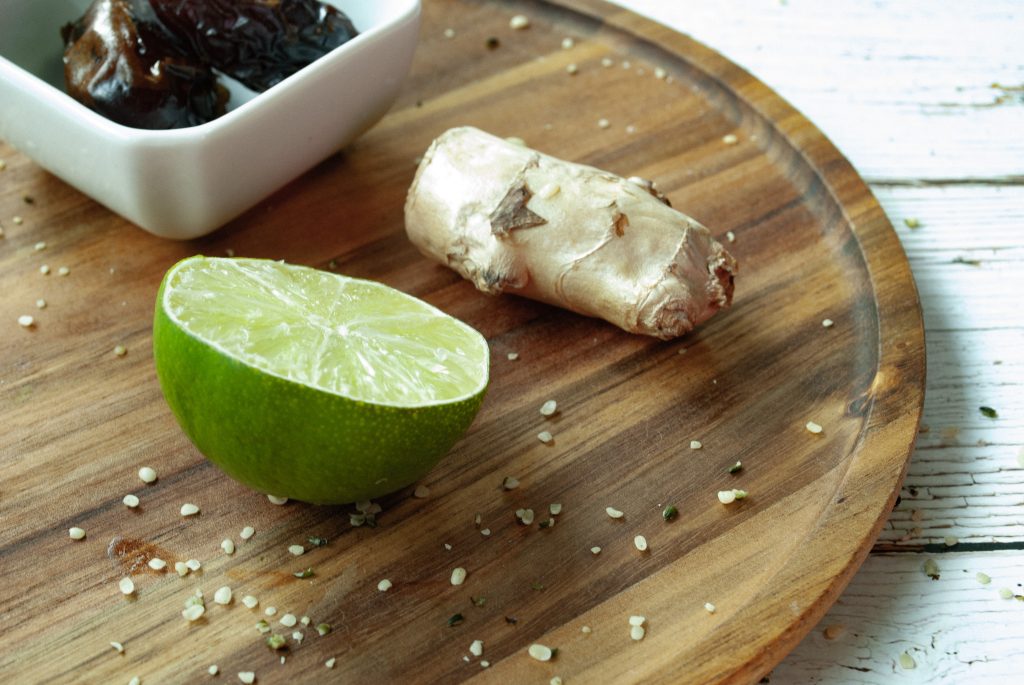 Ginger root, half of a lime and a bowl of dates sitting on a round wooden board.